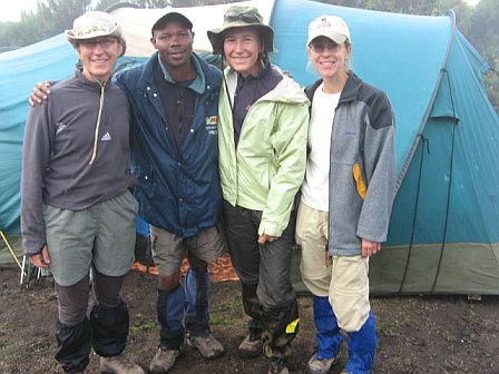 Group of four hikers smiling outdoors near a blue tent, dressed in outdoor gear for trekking.