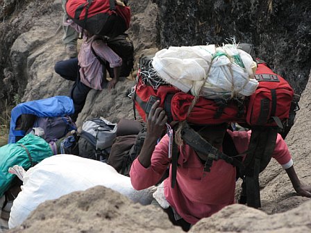 Climbers ascending Kilimanjaro with heavy backpacks and gear on a rugged mountain trail.
