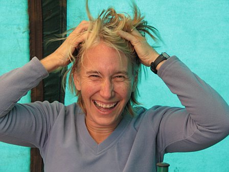 Woman with messy blonde hair laughing and holding her head, outdoor setting with a blue background.