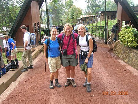 Group of three hikers with backpacks at Kilimanjaro entrance, smiling and ready for the trek, surrounded by lush greenery and other hikers.