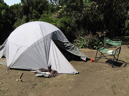 Camping tent set up outdoors with a chair and supplies, surrounded by trees and nature.