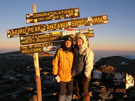 Two climbers at Uhuru Peak on Mount Kilimanjaro, celebrating reaching Africa's highest point at 5895 meters, with a scenic sunset in the background.