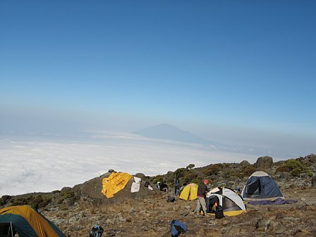 Campers and tents on Mount Kilimanjaro's summit crater with clouds and distant mountain view.