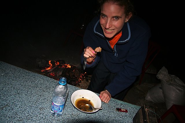 Woman smiling at camera while eating outdoors at night near a fire, with a bowl of food and a water bottle on the table.