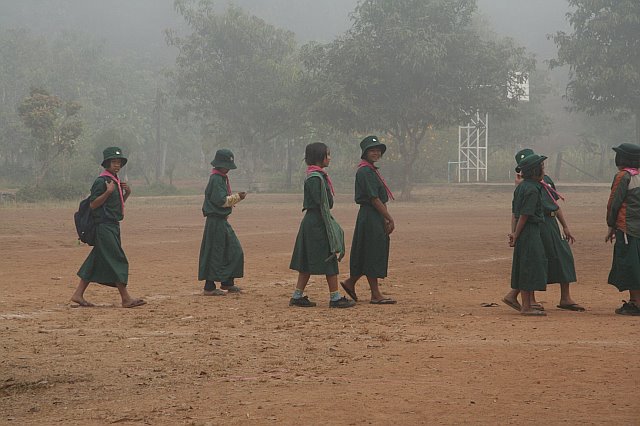 Group of girl scouts in uniform walking in a line on a dusty field with trees and fog in the background during Chiang Mai adventure.