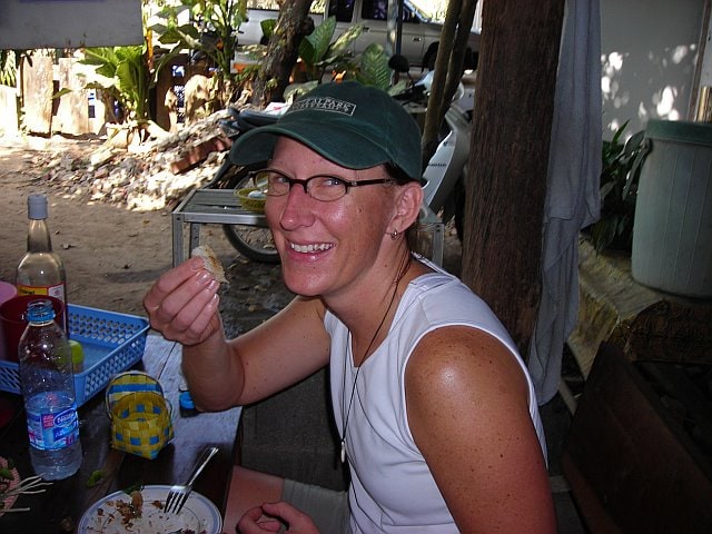 Smiling woman with glasses and a cap, enjoying outdoor dining in Chiang Mai, Thailand, during a travel adventure.
