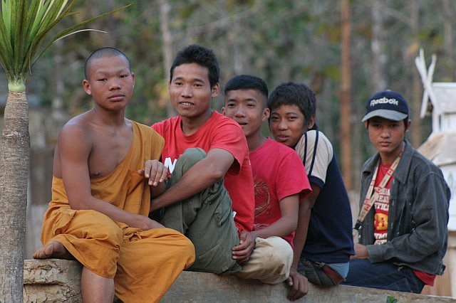 Group of young boys sitting outdoors in Chiang Mai, Thailand, during a nighttime adventure, with a monk and local residents in the background.