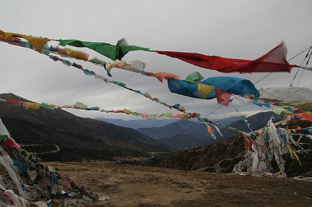 Colorful prayer flags flutter in the wind over a mountainous landscape with rugged terrain and cloudy sky.