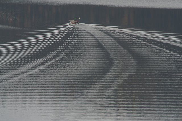 A boat creating ripples on a calm lake during sunset, with a distant shoreline in the background.