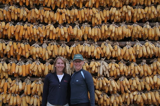 Two women standing in front of a large wall of hanging corn cobs drying in the sun, showcasing traditional agricultural practices.