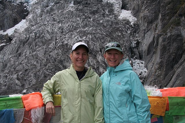 Two women in outdoor jackets smiling in front of a rocky mountain landscape with prayer flags.
