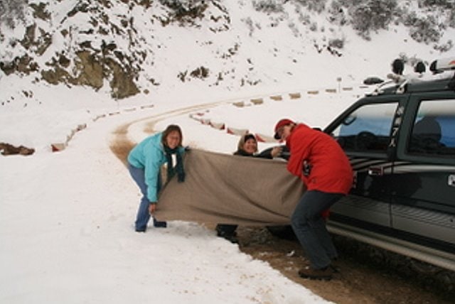 Group of people in winter gear setting up a blanket near a black vehicle on snowy mountain road.