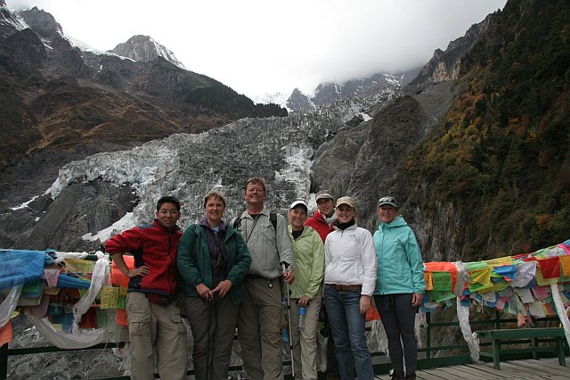 Group of hikers standing on a bridge with colorful prayer flags, mountain landscape with glacier and snow-capped peaks in the background.