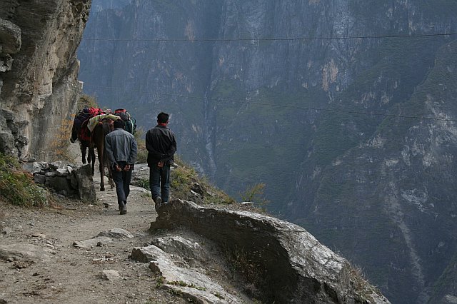 Hikers walking along a narrow trail with a steep cliff on one side and a deep gorge below, surrounded by rugged mountain terrain.