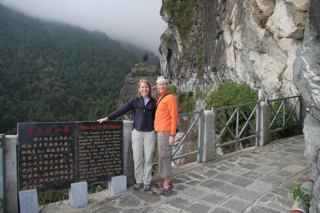 Two women standing on a mountain trail with a scenic view of misty mountains and lush greenery in the background.