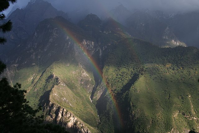 Scenic view of Tiger Leaping Gorge with a vibrant rainbow arching over lush green mountains and rugged cliffs in China.