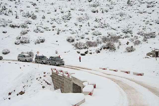 Snow-covered mountain road with parked cars and a person walking in winter landscape.