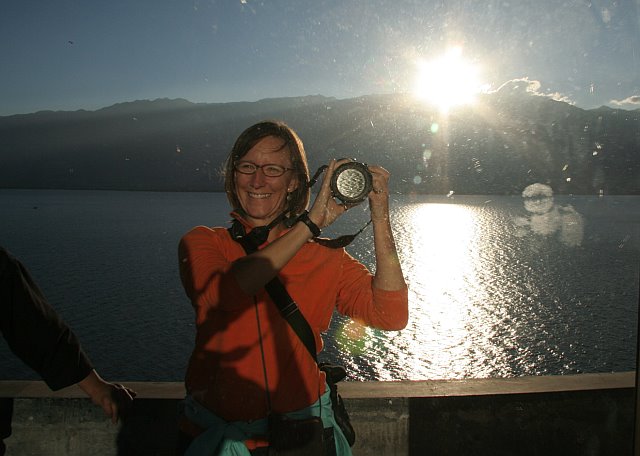 Smiling woman taking a photo with a camera on a boat during sunset over a lake with mountains in the background.