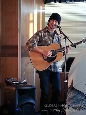 A man wearing a black beanie and plaid shirt smiles while playing an acoustic guitar and singing into a microphone inside an Antarctic expedition ship lounge. A speaker sits beside him as he performs for passengers in the warm wood paneled room.