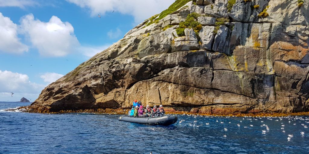 A group of people in colorful outdoor gear rides in a black inflatable boat along a rocky coastline with towering cliffs streaked with moss and lichen. The clear blue sky and scattered seabirds overhead enhance the sense of exploration in this remote, oceanic setting.