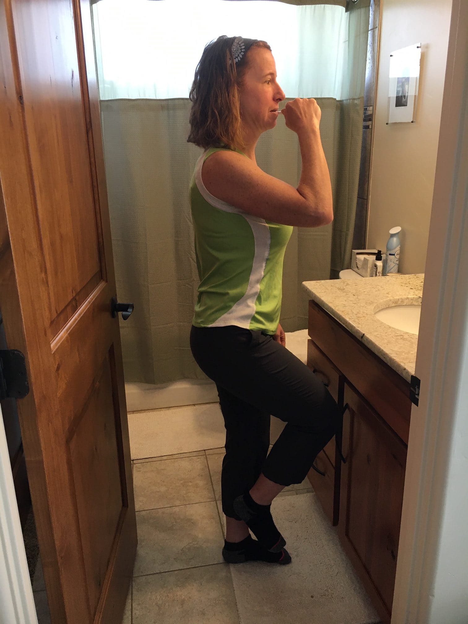 A woman in a green sleeveless top and dark workout pants balances on one foot while brushing her teeth in a bathroom. She looks into the mirror with focus, practicing stability as part of her routine. The cozy bathroom features a wooden vanity, a green shower curtain, and a well-lit, relaxed atmosphere.