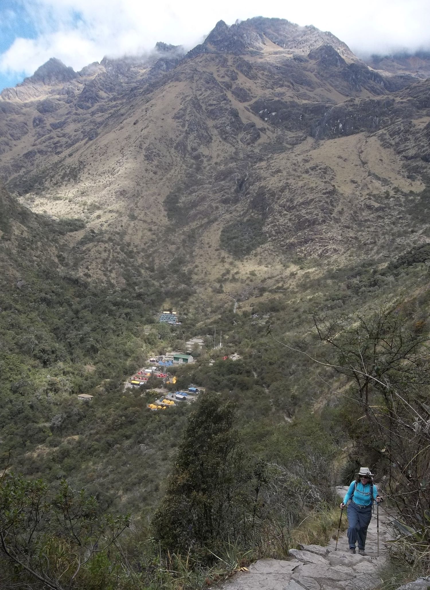 A hiker wearing a sun hat, sunglasses, and a blue jacket ascends a rugged stone path with trekking poles, surrounded by towering mountains. Below, a small campsite with colorful tents and buildings is nestled in the valley, contrasting against the steep, rocky slopes. The mist-shrouded peaks and vast landscape create a dramatic and awe-inspiring scene of high-altitude trekking.