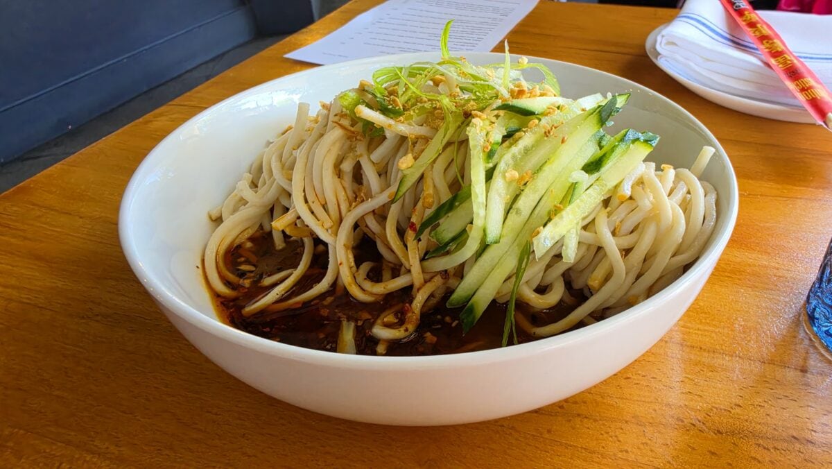 Bowl of Asian-style noodles with cucumber and green onion garnish on a wooden table.