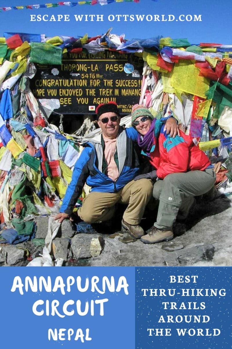 Two hikers at the Annapurna Circuit in Nepal, smiling and sitting under colorful prayer flags at Thorong La Pass, celebrating reaching 5,416 meters during their trek.