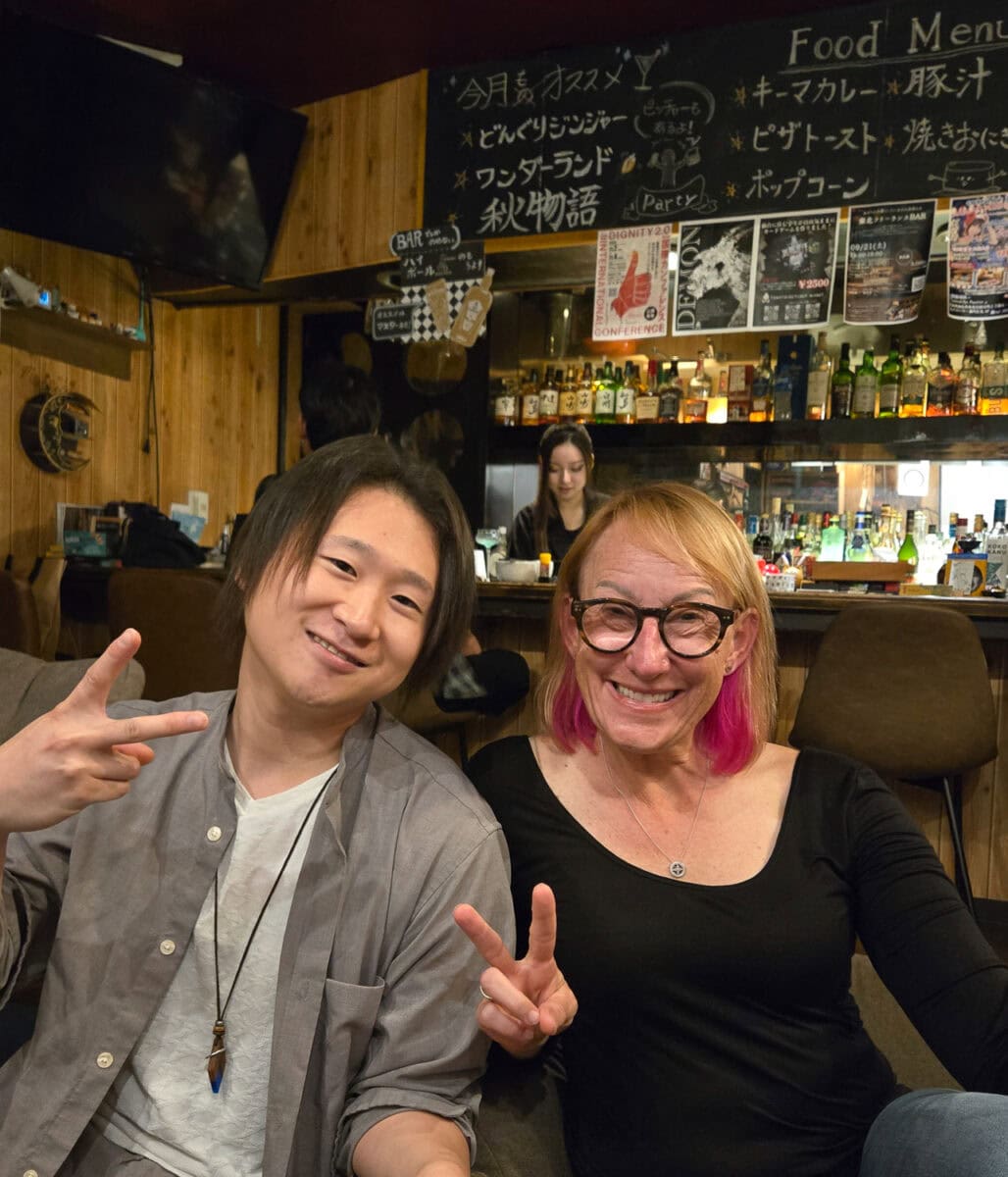 Smiling woman with pink hair and glasses and young man making peace signs at a cozy bar or restaurant.