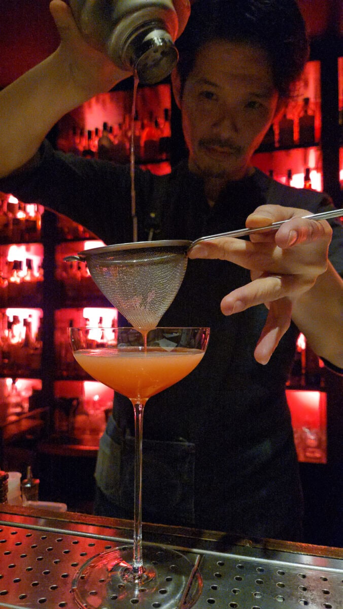 Bartender pouring a cocktail through a strainer into a glass at OttsWorld bar, with a red-lit background.
