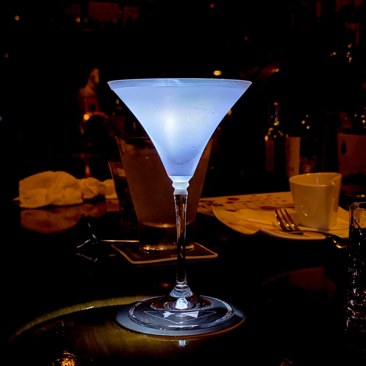 Blue cocktail in a martini glass illuminated in a dimly lit bar setting.