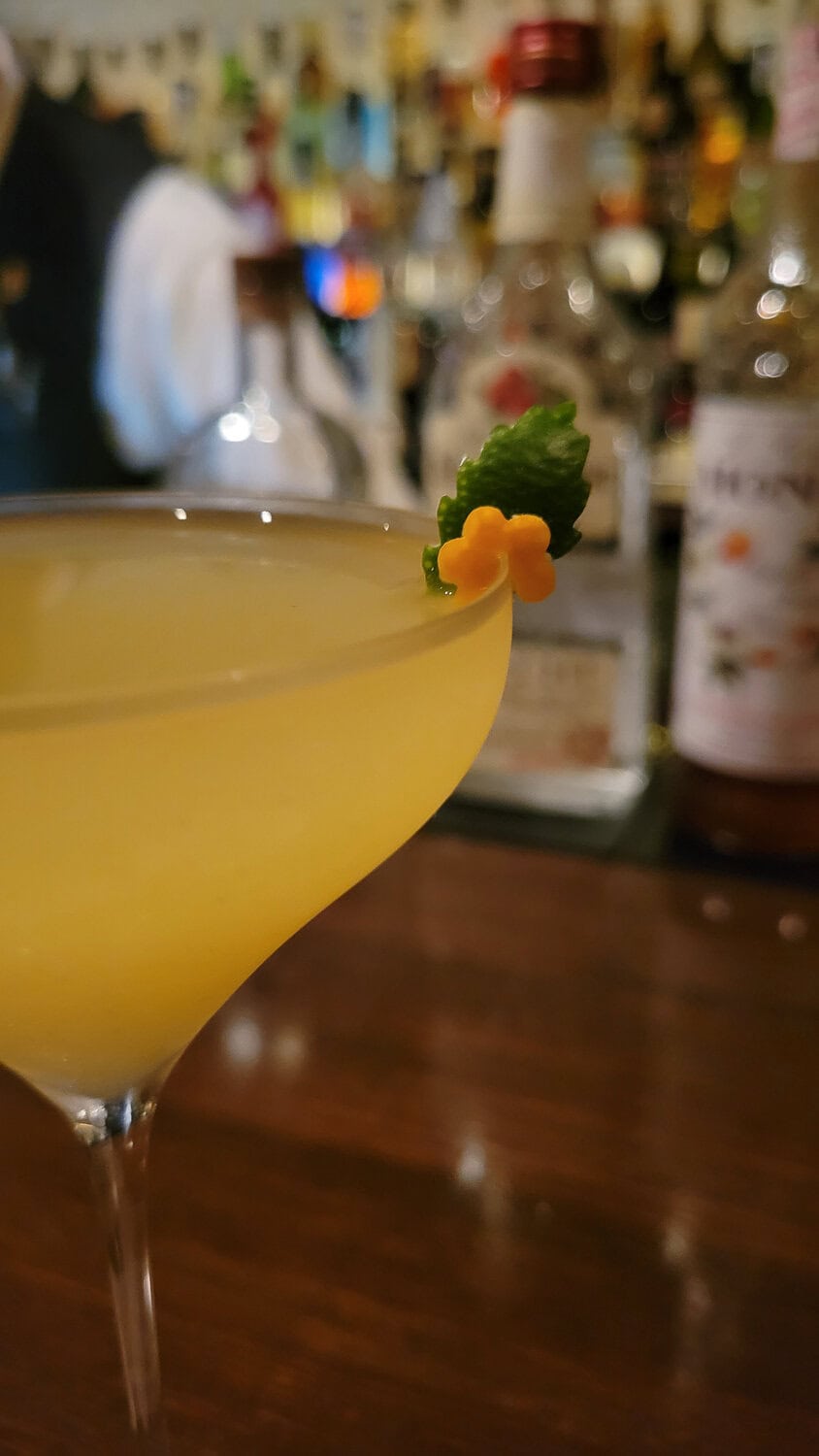 Close-up of a yellow cocktail garnished with a small green leaf and orange flower, with a blurred bar background.