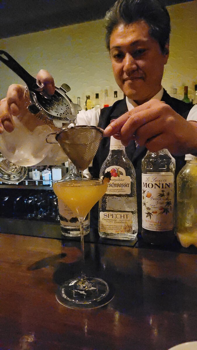 Bartender pouring a cocktail through a strainer into a glass at a bar, with liquor bottles in the background.