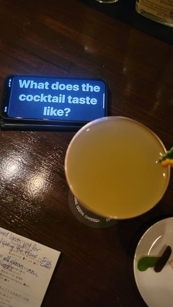 Smartphone displaying the question 'What does the cocktail taste like?' next to a cocktail on a dark wooden table.