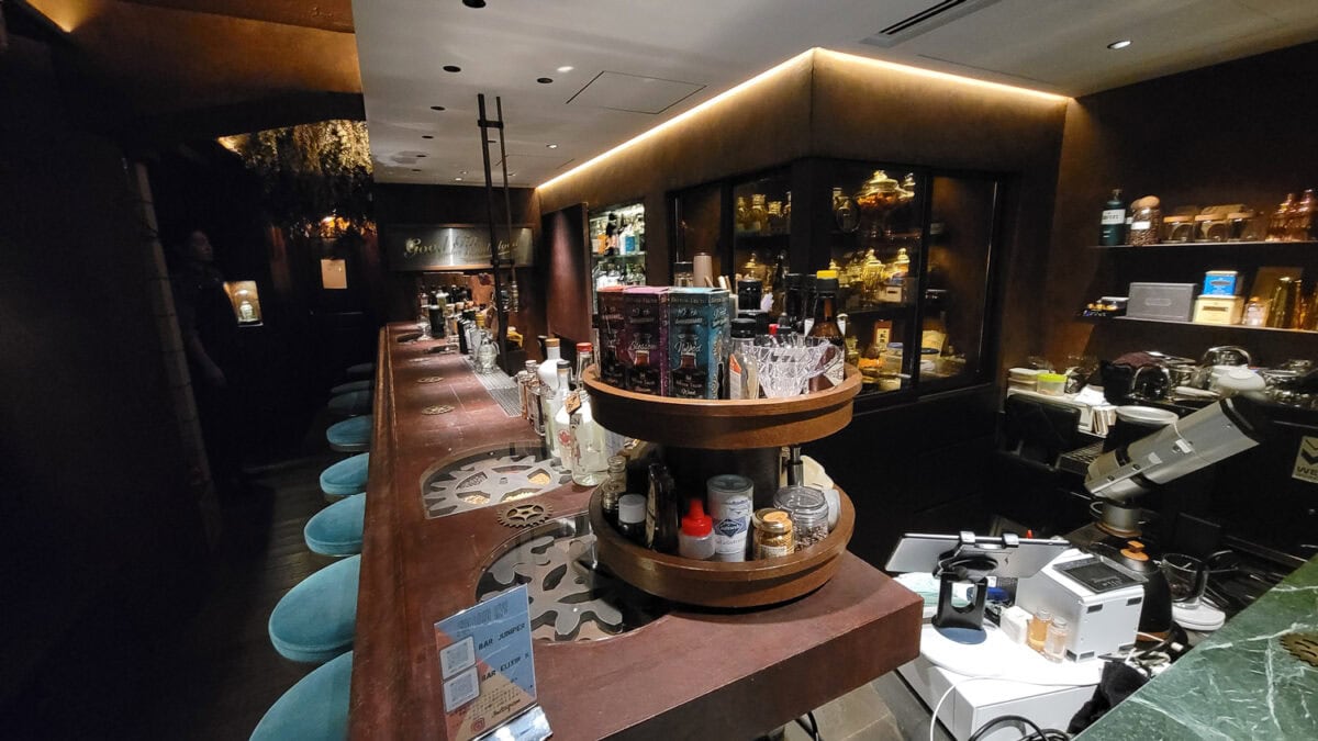 Modern bar interior with a long wooden counter, blue bar stools, and a well-stocked display of bottles and glassware in a stylish, dimly lit setting.