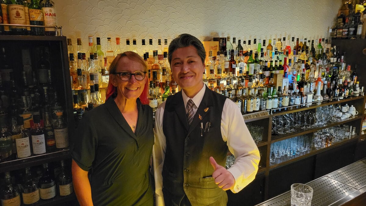 Smiling bartender in a black vest and white shirt giving a thumbs-up behind a well-stocked bar with various liquor bottles.