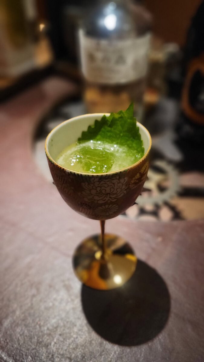 Close-up of a green cocktail garnished with a fresh mint leaf in an ornate glass on a bar counter.