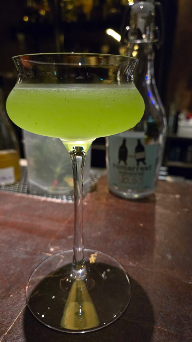 Green cocktail in a coupe glass on a bar counter with a bottle in the background.