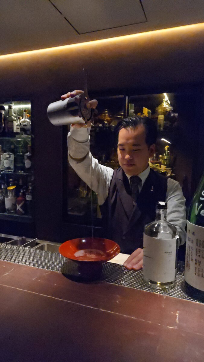 Bartender pouring a drink at a bar with bottles and glasses in the background, creating a lively nightlife scene.