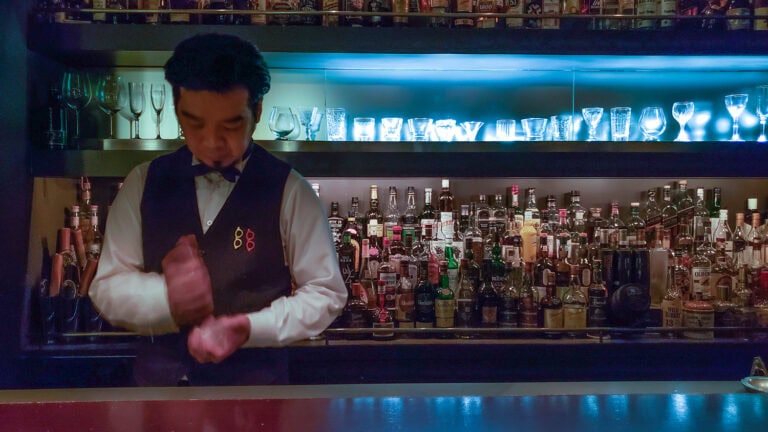 Bartender preparing a cocktail at a well-stocked bar with illuminated bottles in the background.