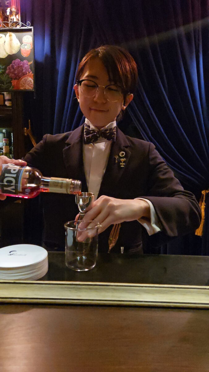 Bartender in formal attire pouring a drink at a bar with dark curtains and decorative lighting in the background.
