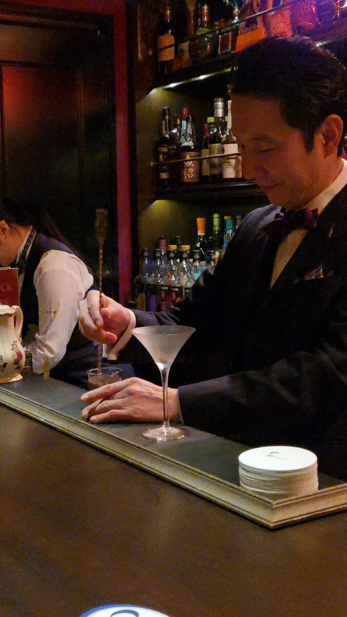 Elegant bartender in a tuxedo preparing a cocktail at a sophisticated bar with liquor bottles in the background.