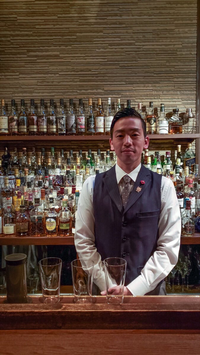 Bartender standing behind a bar with a variety of liquor bottles in the background, wearing a vest and white shirt.
