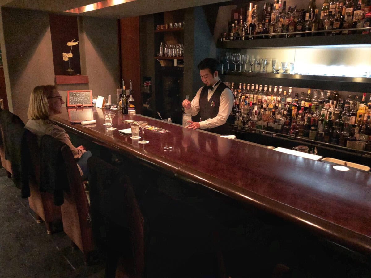 A bartender serving drinks at a dimly lit bar with two customers seated at the counter.