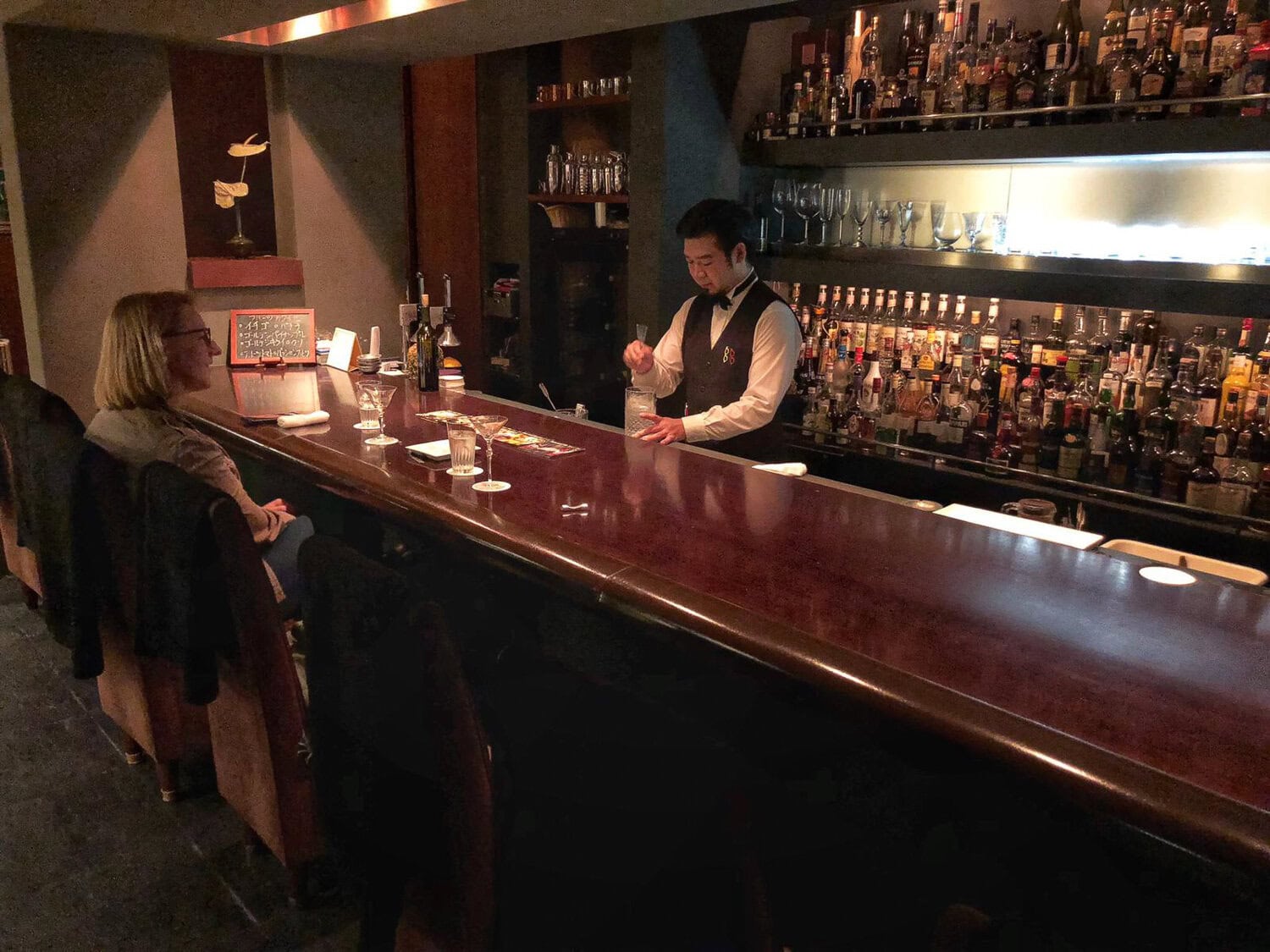 A bartender serving drinks at a dimly lit bar with two customers seated at the counter.