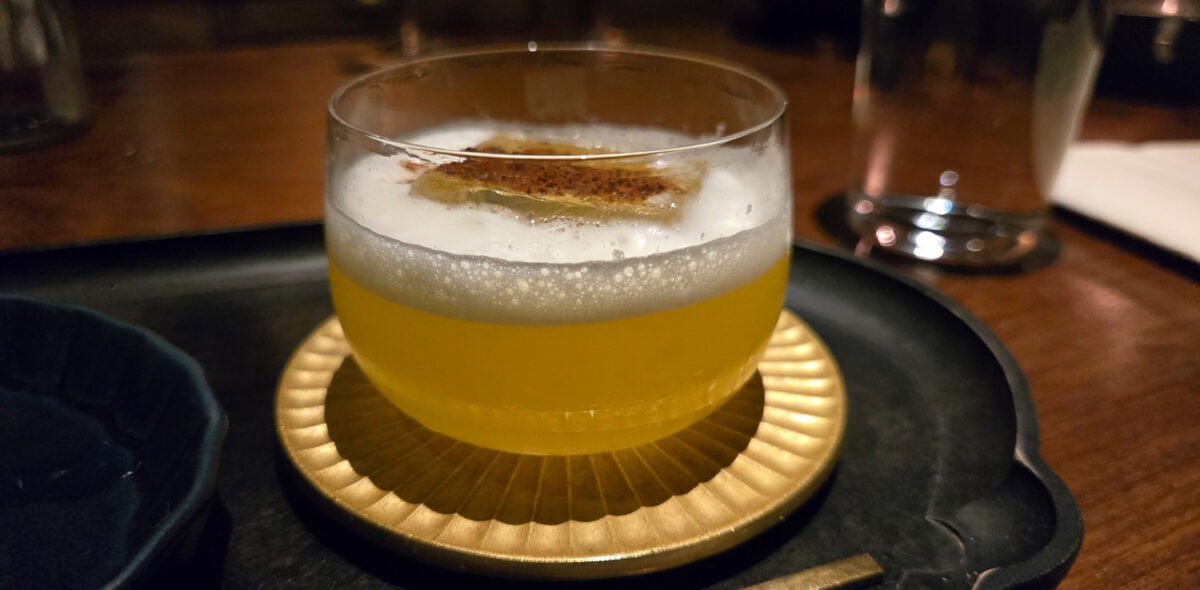 Close-up of a cocktail with foam and a cinnamon stick garnish on a black tray.