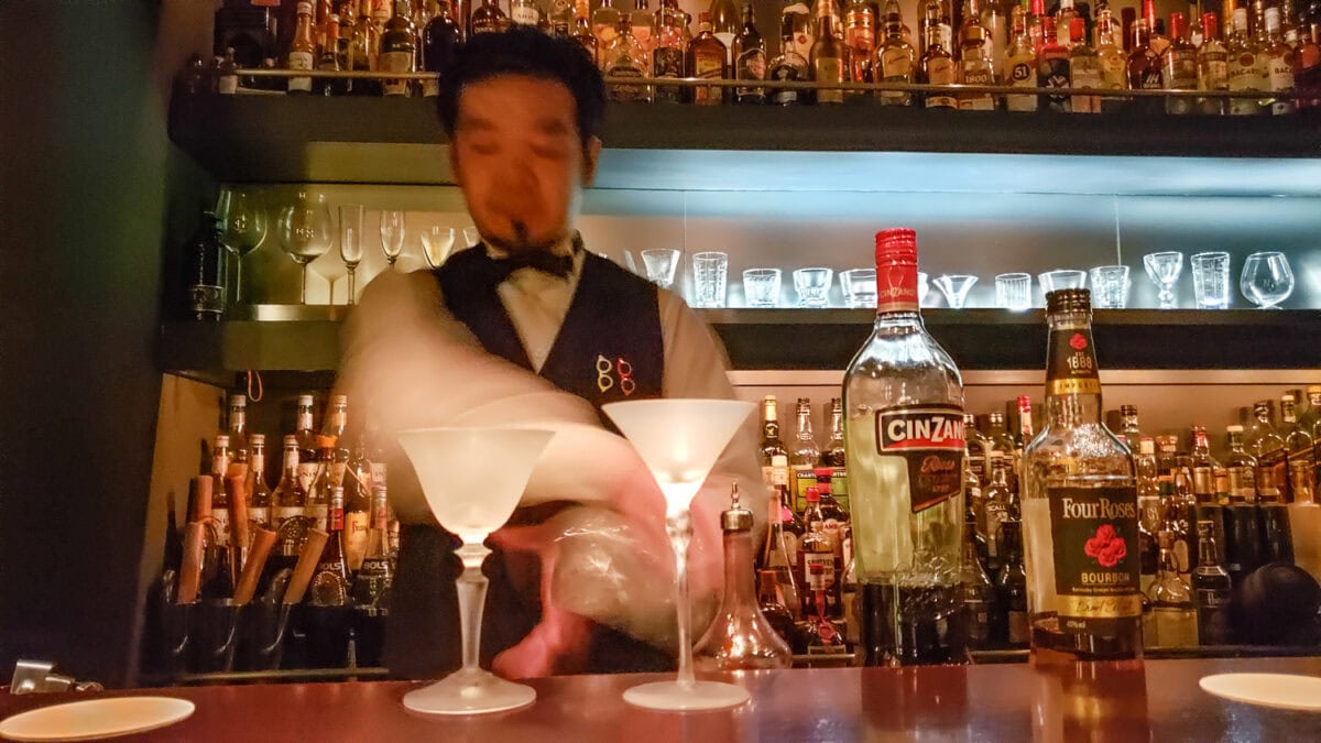 Bartender preparing cocktails at a bar with various liquor bottles in the background.