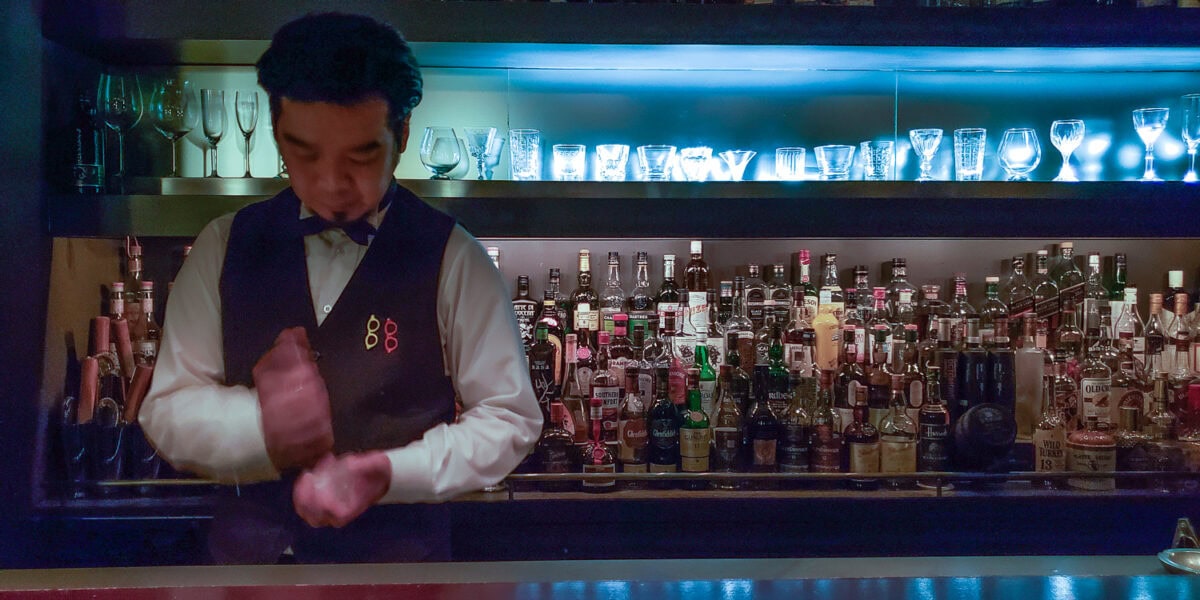 Bartender preparing a cocktail at a well-stocked bar with colorful bottles and glassware.