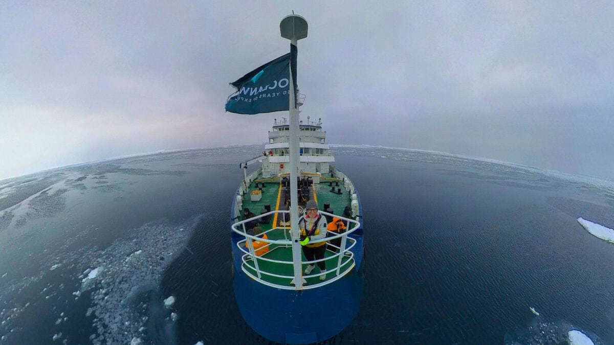 Research vessel navigating icy waters in polar region with crew onboard, under cloudy sky.