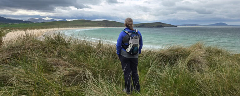 Hiker standing on grassy dunes overlooking a scenic beach with turquoise waters and distant hills.